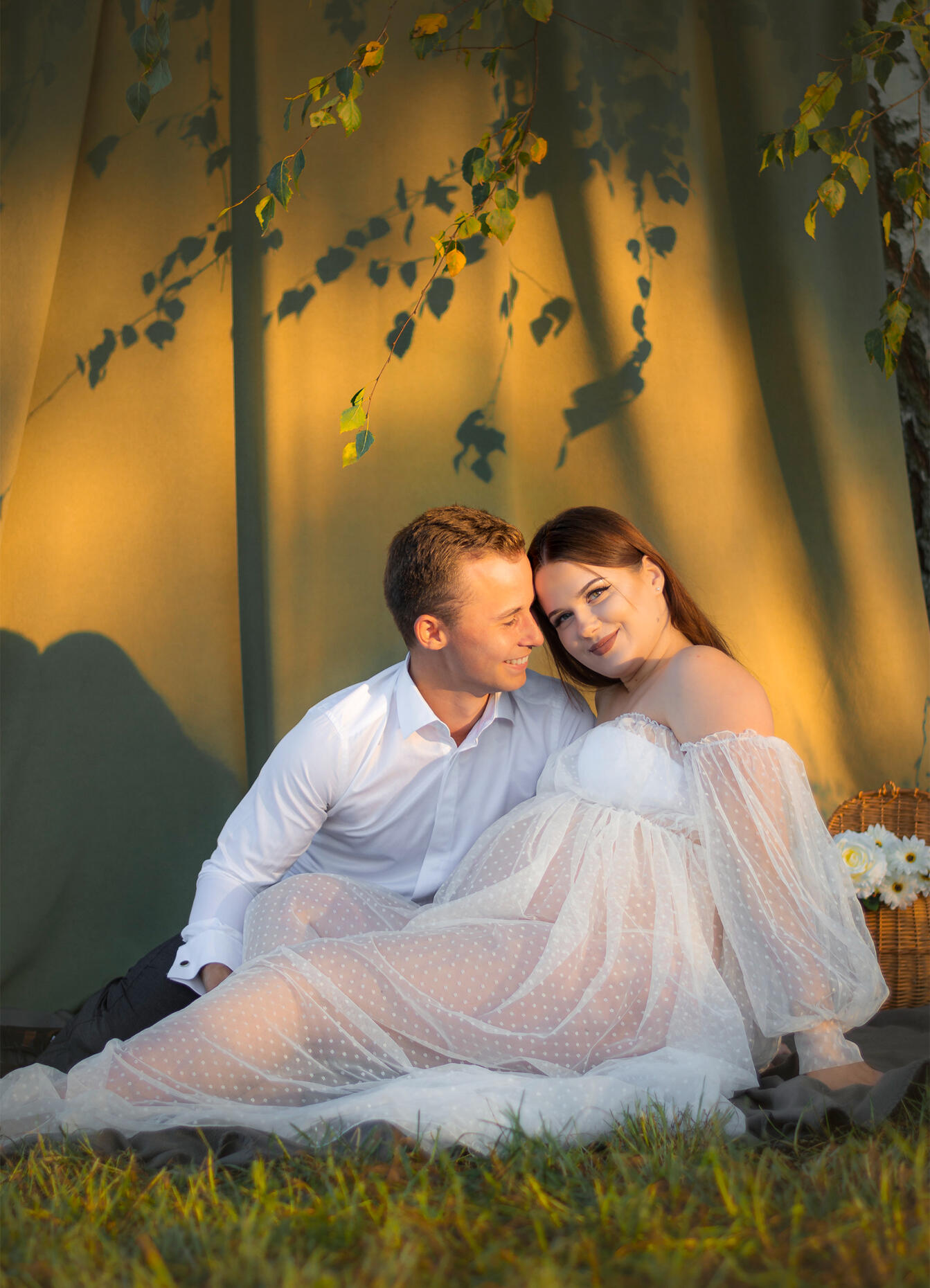 Untitled nėščia moteris su partneriu laimingi sėdi tarp žaliuojančių beržo šakų, į juos šviečia vakarėjanti saulė, jie pozuoja